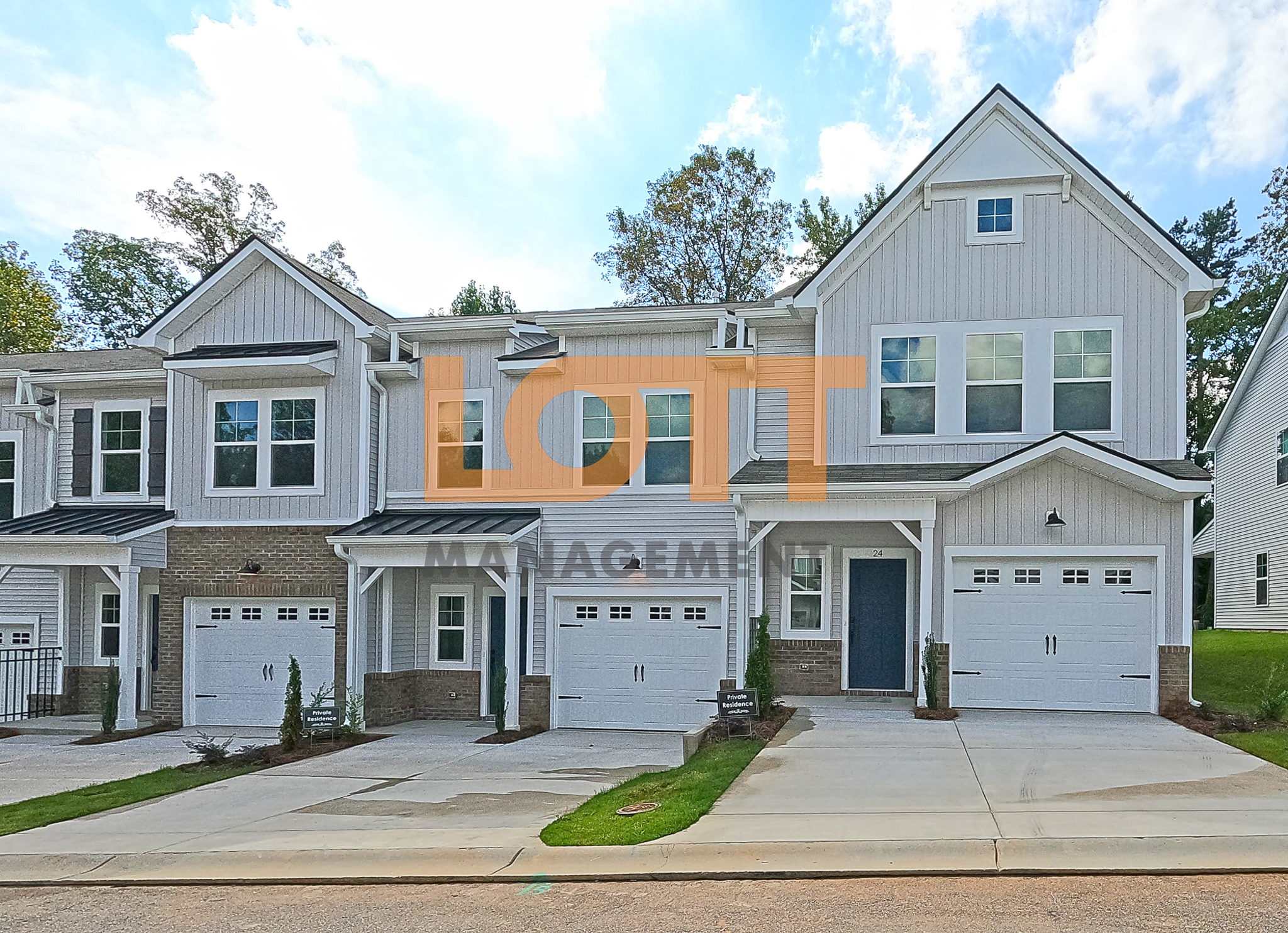 A front-facing photograph of a row of modern townhouses with light gray vertical siding, white trim, and dark gray roofs. Each unit features a two-car white garage door with black decorative handles, a dark blue front door, and a tan brick base near the entrance. The rightmost unit is marked with the house number 