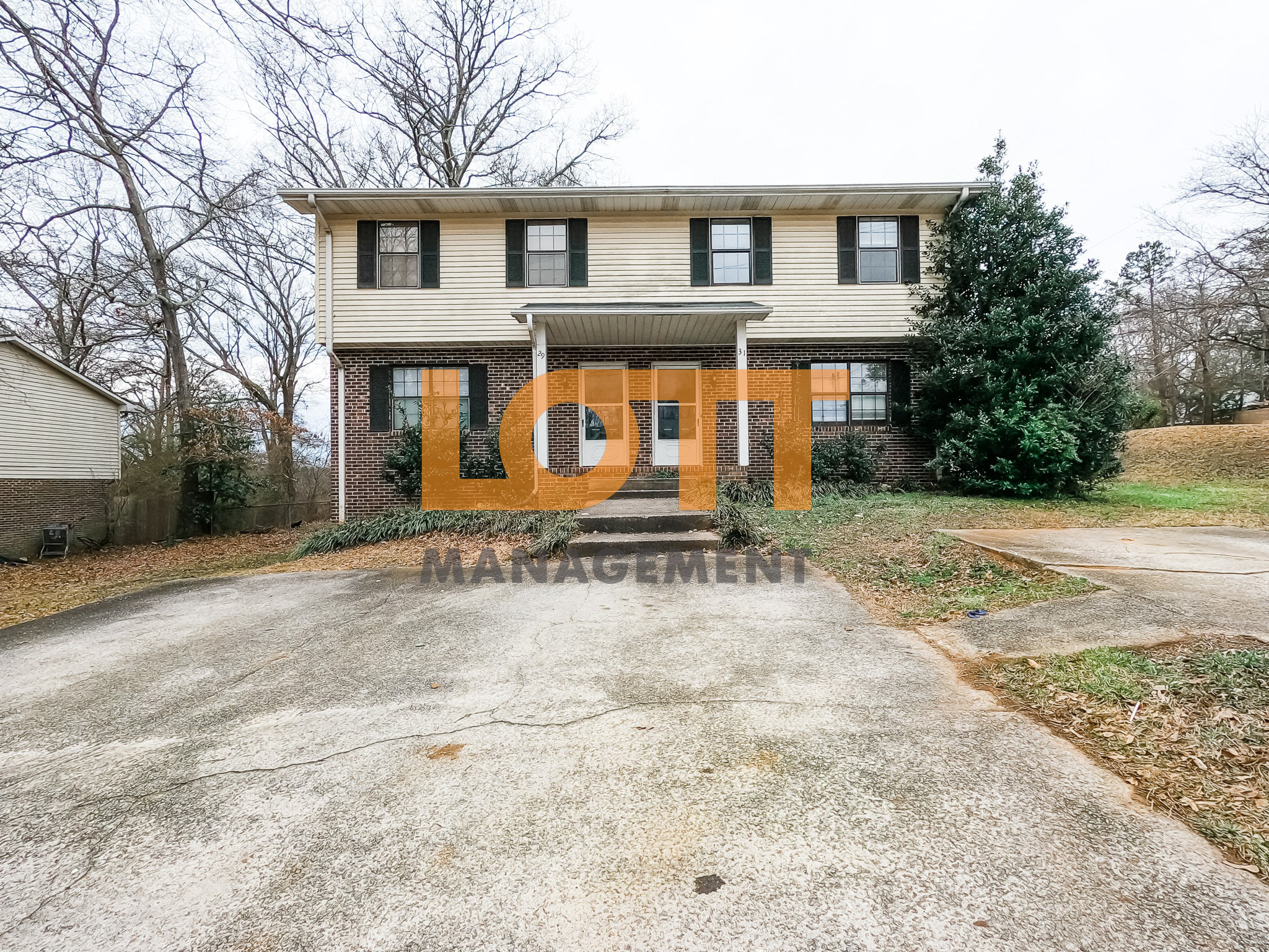 Front view of a two-story duplex with a brick lower level and light beige siding on the upper level. The building features two white front doors marked '29' and '31', each under a small portico with white columns. Eight windows with dark shutters are evenly spaced across the facade. A large concrete driveway with visible cracks leads to the house, bordered by patchy grass and small shrubs. A dense evergreen tree stands to the right, while bare deciduous trees are visible in the background under an overcast sky. A semi-transparent 'LOTT MANAGEMENT' watermark is overlaid on the image.