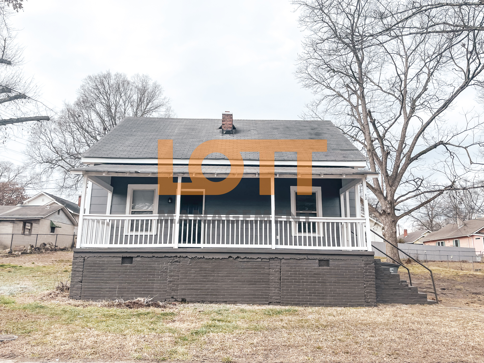 Front view of a single-story dark blue house with a white porch, railing, and square columns, set on a dark grey brick foundation. The house features a gabled roof, a central brick chimney, and two white-framed windows flanking a dark front door. A patchy lawn with dry grass and bare dirt surrounds the house, with large leafless trees and neighboring properties visible in the background under an overcast sky. A semi-transparent 'LOTT MANAGEMENT' watermark overlays the image.
