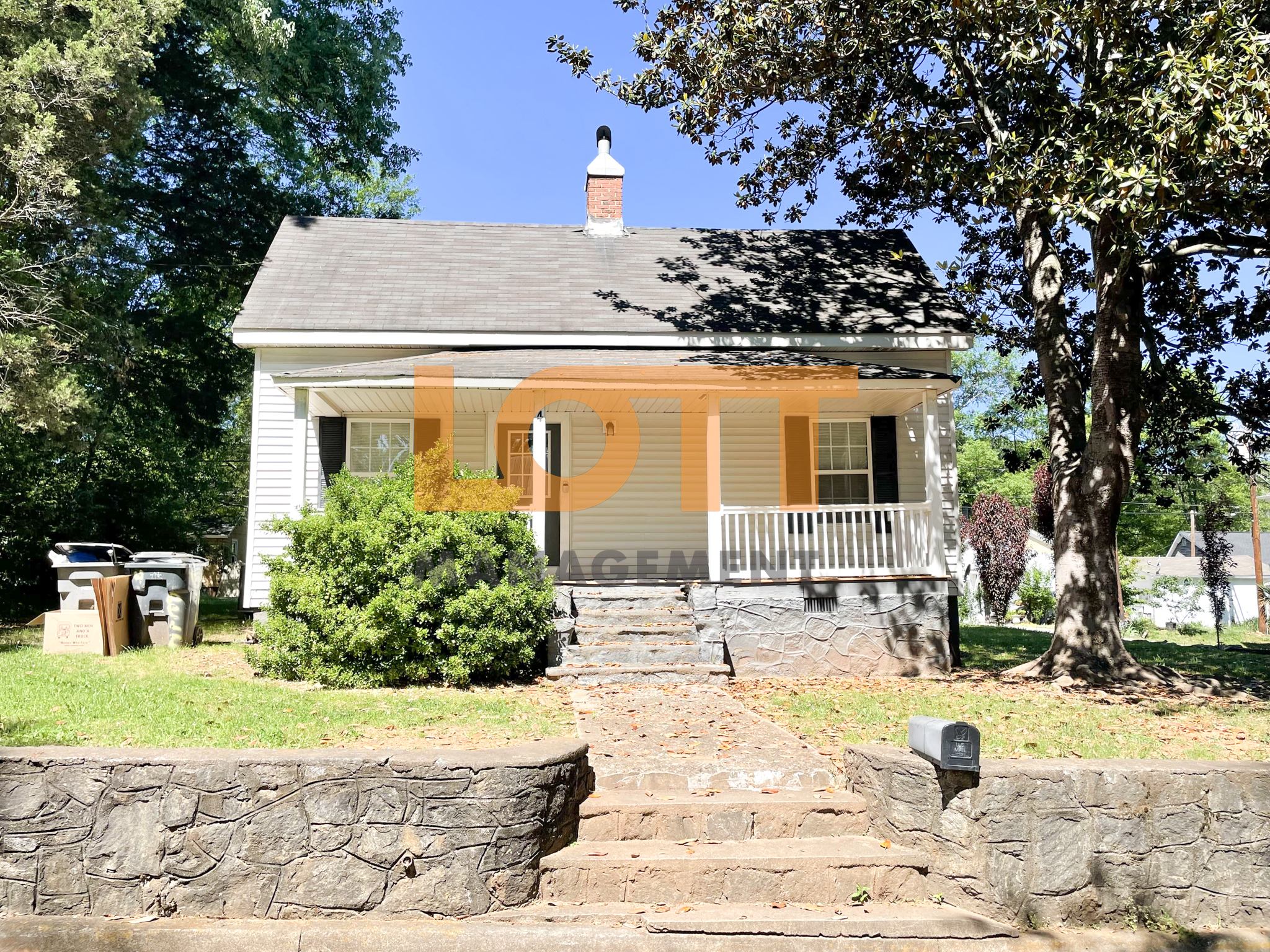 A front-facing photograph of a single-story white house with horizontal siding, a dark gray shingled gable roof, and a red brick chimney. The house features a full-width covered front porch supported by white square columns, with a dark front door and two white-framed windows with black shutters. The foundation and retaining walls are covered in a gray stone-like facade. A stone pathway with steps leads to the porch, flanked by green grass and a large leafy bush on the left. A mature tree with dense foliage stands on the right, casting shadows over the yard and house. The foreground includes a gray mailbox on the retaining wall and trash bins with cardboard boxes on the far left. The background shows a clear blue sky and additional trees. A semi-transparent orange 