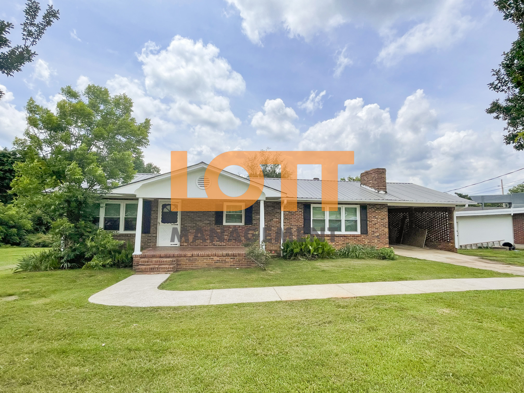 A front-facing photograph of a single-story reddish-brown brick house with a gray metal gabled roof and a white circular vent on the front gable. The house features a recessed front porch with a white column, a white front door accessed by three brick steps, and multiple windows with white frames and dark shutters. To the right, an attached brick carport with an open side reveals stored items, and a white garage or shed with a garage door is visible beyond it. The foreground includes a lush green lawn, a curved concrete sidewalk leading to the porch, and a driveway extending to the carport. The background features trees and shrubs under a blue sky with scattered white clouds. A semi-transparent orange 