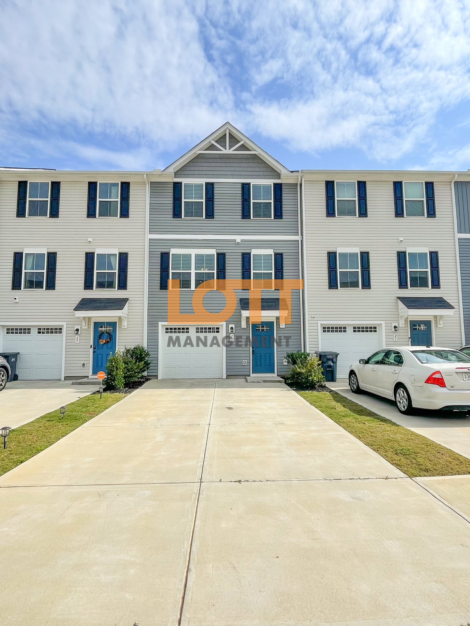 A front-facing photograph of three attached two-story townhouses with light beige and grey siding, dark blue shutters, and blue front doors. Each unit features a white garage door, concrete driveways, and small landscaped areas with grass and bushes. The central unit has a distinctive white-trimmed gable above its second-story windows. A white sedan is parked in the driveway of the rightmost unit, and trash cans are visible near the left and right driveways. The background includes a bright blue sky with scattered white clouds. A semi-transparent 