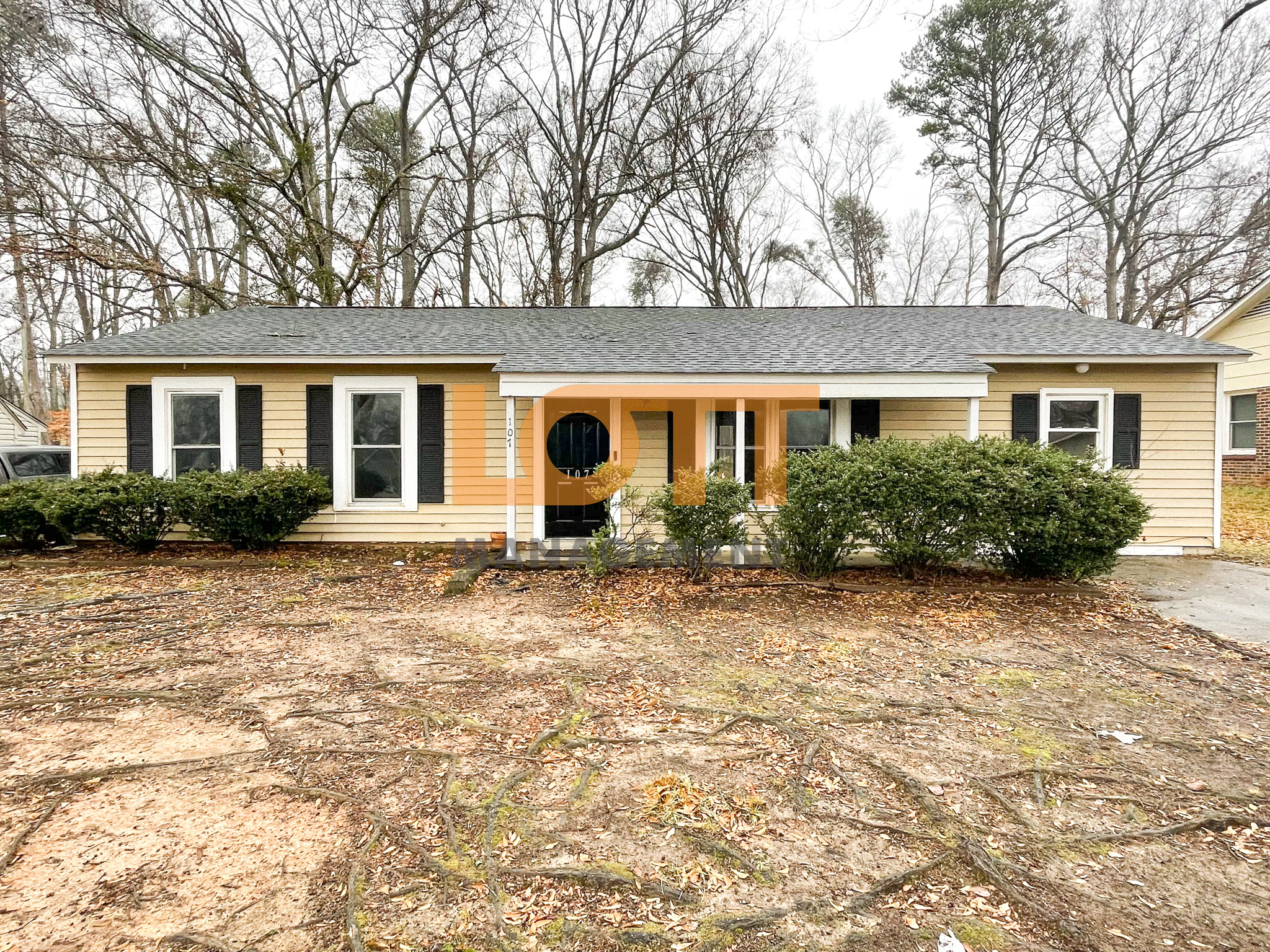 A front-facing photograph of a single-story light yellow ranch-style house with a dark gray shingled roof and black-shuttered windows. The house features a central covered porch with white pillars, a black front door with a circular glass design, and the house number 