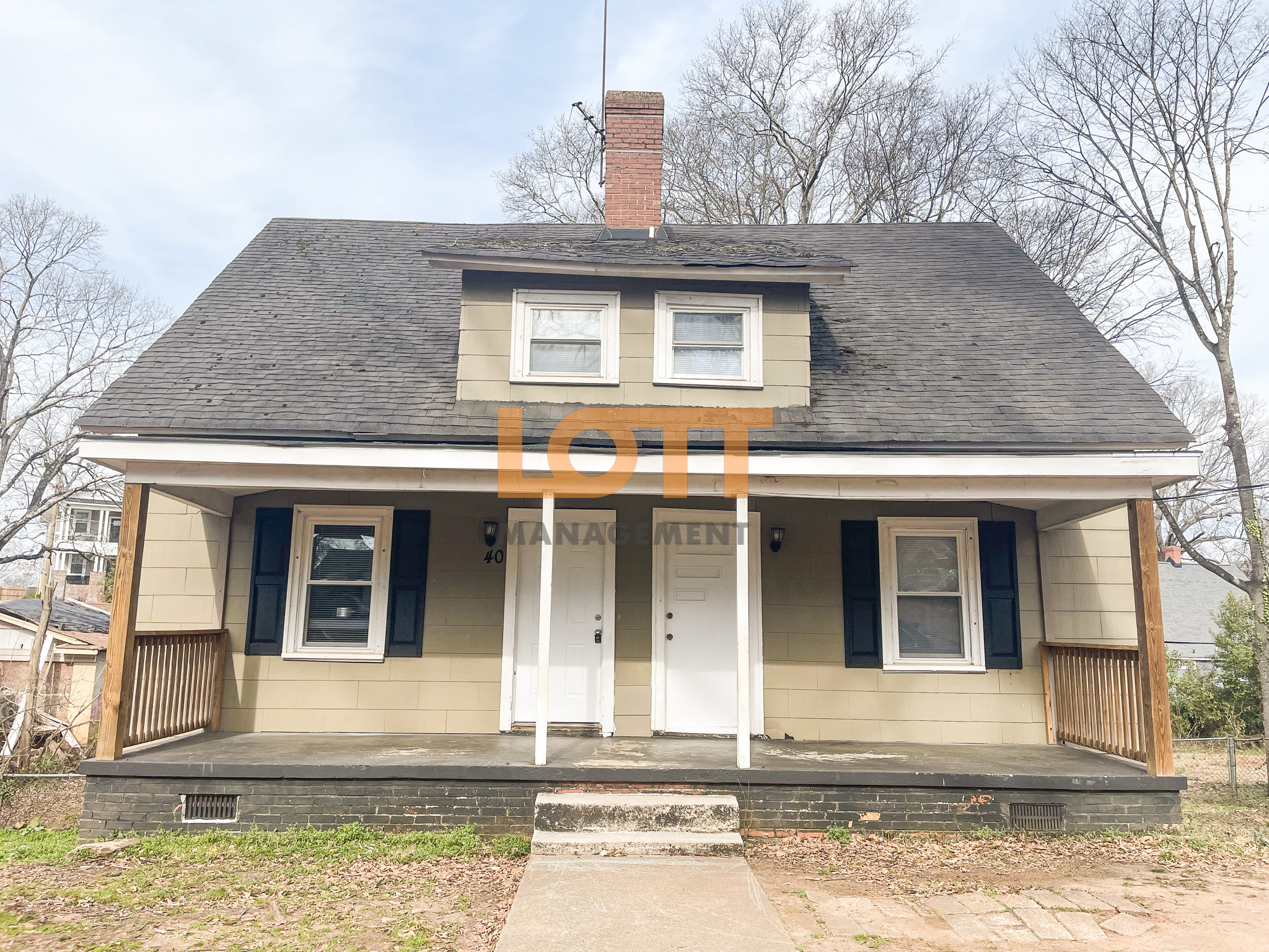 Two-story light tan duplex with a dark gray roof, central dormer, and a full-width front porch supported by white columns. Two white front doors and black-shuttered windows are visible, with a sparse lawn and bare trees in the background under an overcast sky. 'LOTT MANAGEMENT' watermark is overlaid.