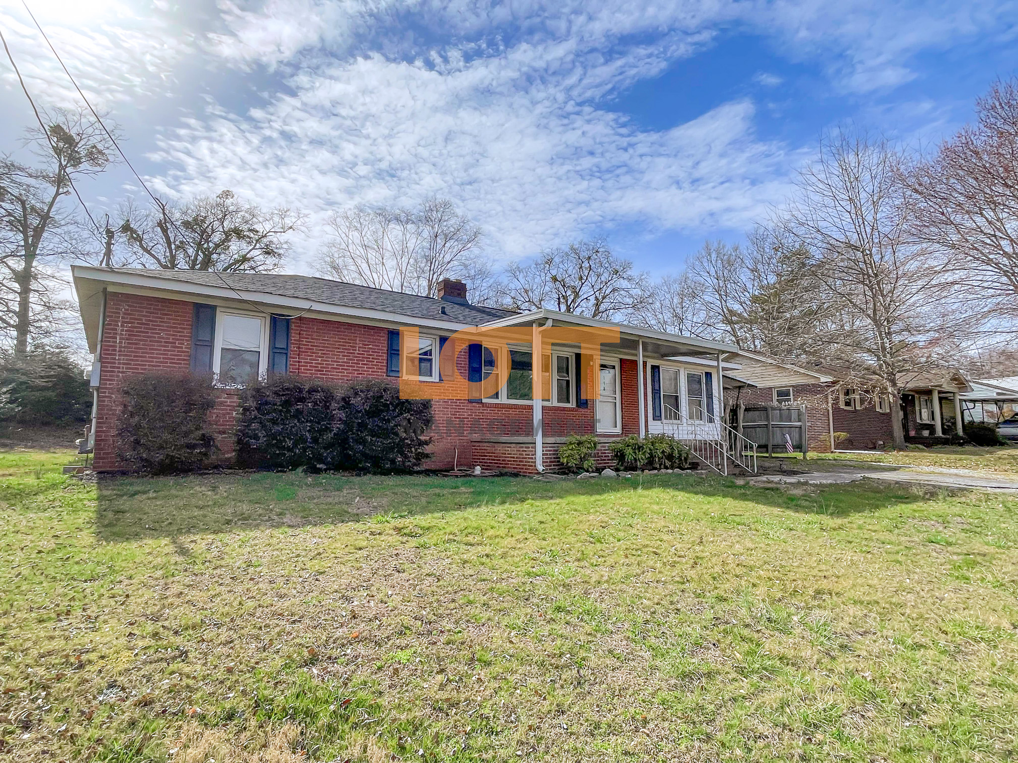 Single-story red brick house with dark blue shutters, a dark gray roof, and a covered front porch/carport supported by white columns. A grassy lawn with green and brown patches surrounds the house, with bare trees and a partly cloudy blue sky in the background. A large 'LOTT MANAGEMENT' watermark is overlaid across the house.