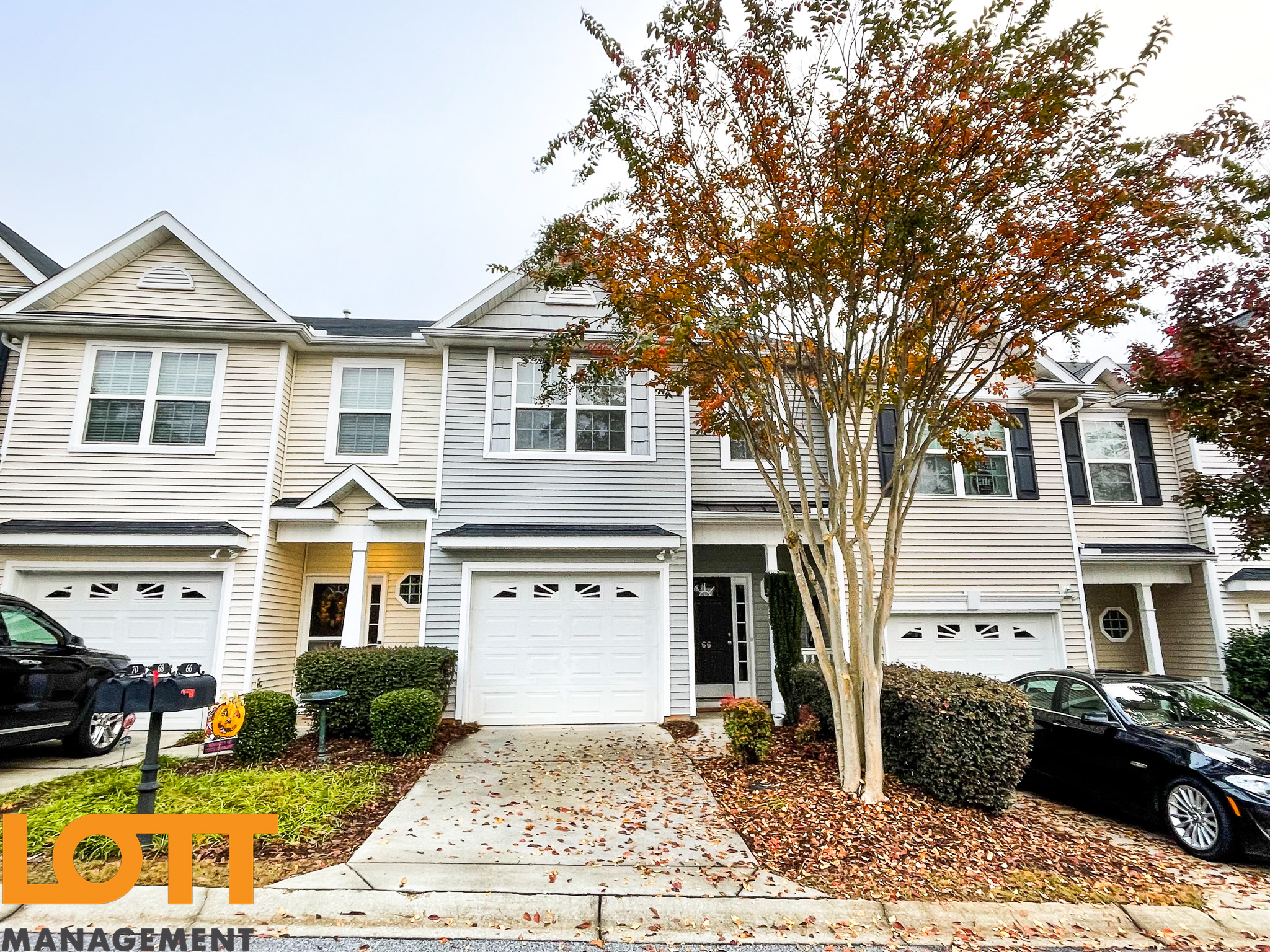 Front view of a row of two-story townhouses with beige and light grey siding, white garage doors, and black-framed windows. The central townhouse features the number '66' on its black front door. A large tree with autumn foliage stands in front of the middle units, and the ground is covered with fallen leaves. Two black cars are parked in the driveways, and a cluster of black mailboxes with numbers '70, 68, 66' is visible near the leftmost unit. A semi-transparent orange 'LOTT MANAGEMENT' watermark is in the bottom-left corner.