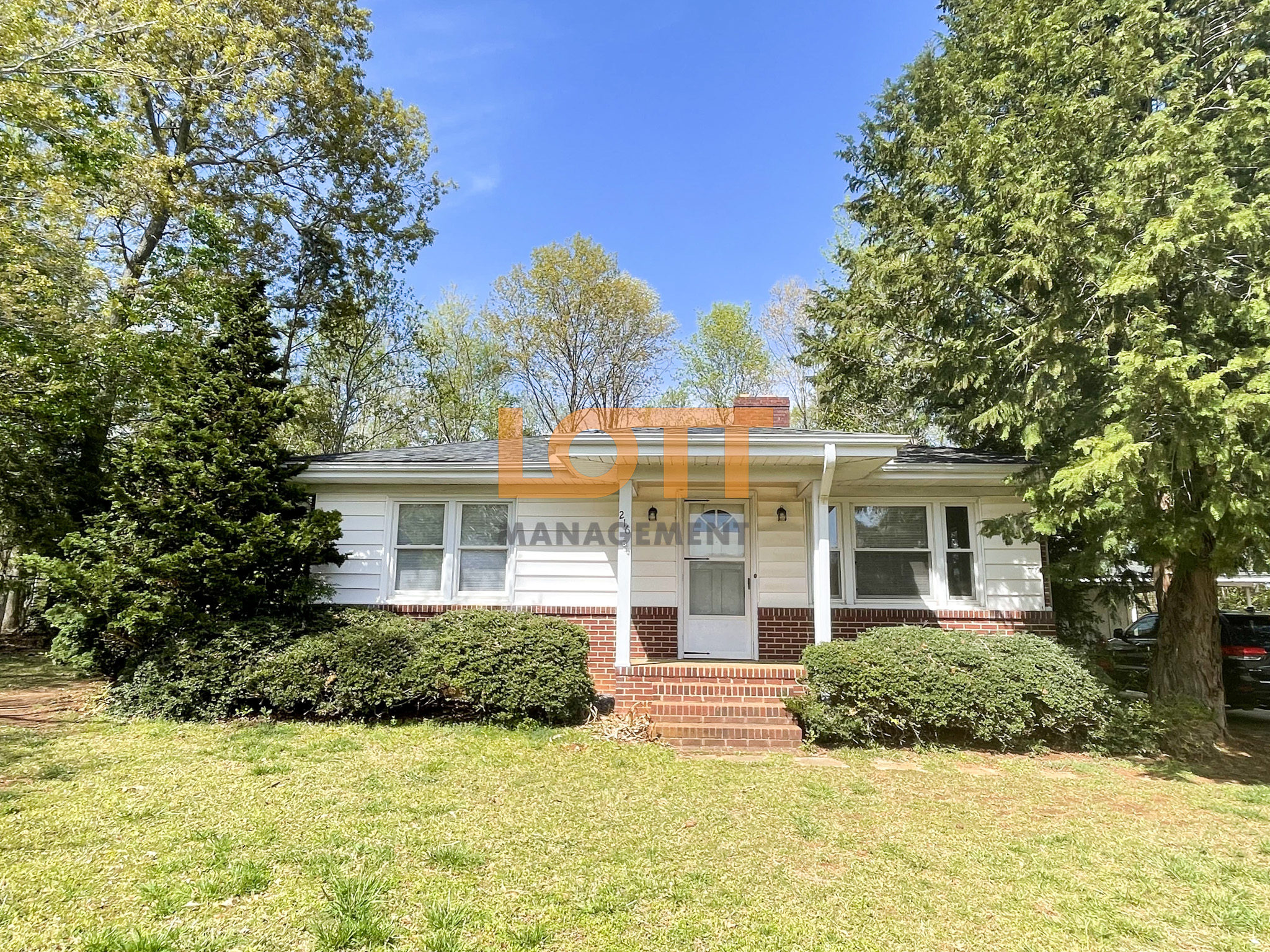 Front view of a single-story white house with horizontal siding, a dark gray shingle roof, and a red brick foundation. The house features a covered porch with white columns, red brick steps, a white front door with an arched window, and black wall-mounted light fixtures. White-framed windows with blinds flank the door, and dense green bushes line the foundation. The yard includes a green lawn, surrounded by mature trees under a clear blue sky. A watermark reading 'LOTT MANAGEMENT' overlays the image, with the number '2' visible above the door.