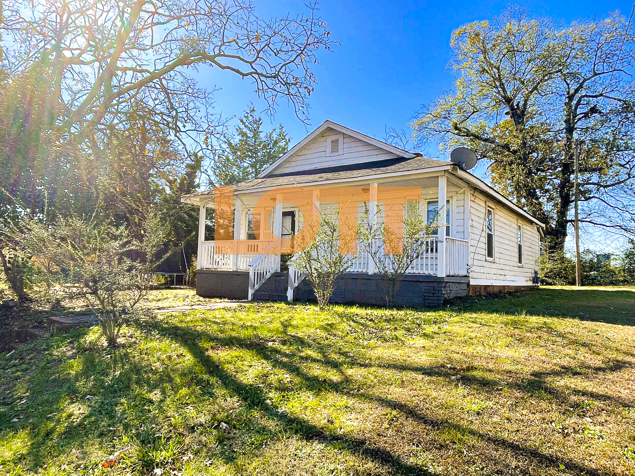 Bright, sunny view of a single-story white house with a front porch, dark gable roof, and a grassy yard surrounded by trees under a clear blue sky. A large orange 'LOTT MANAGEMENT' watermark is overlaid on the house.