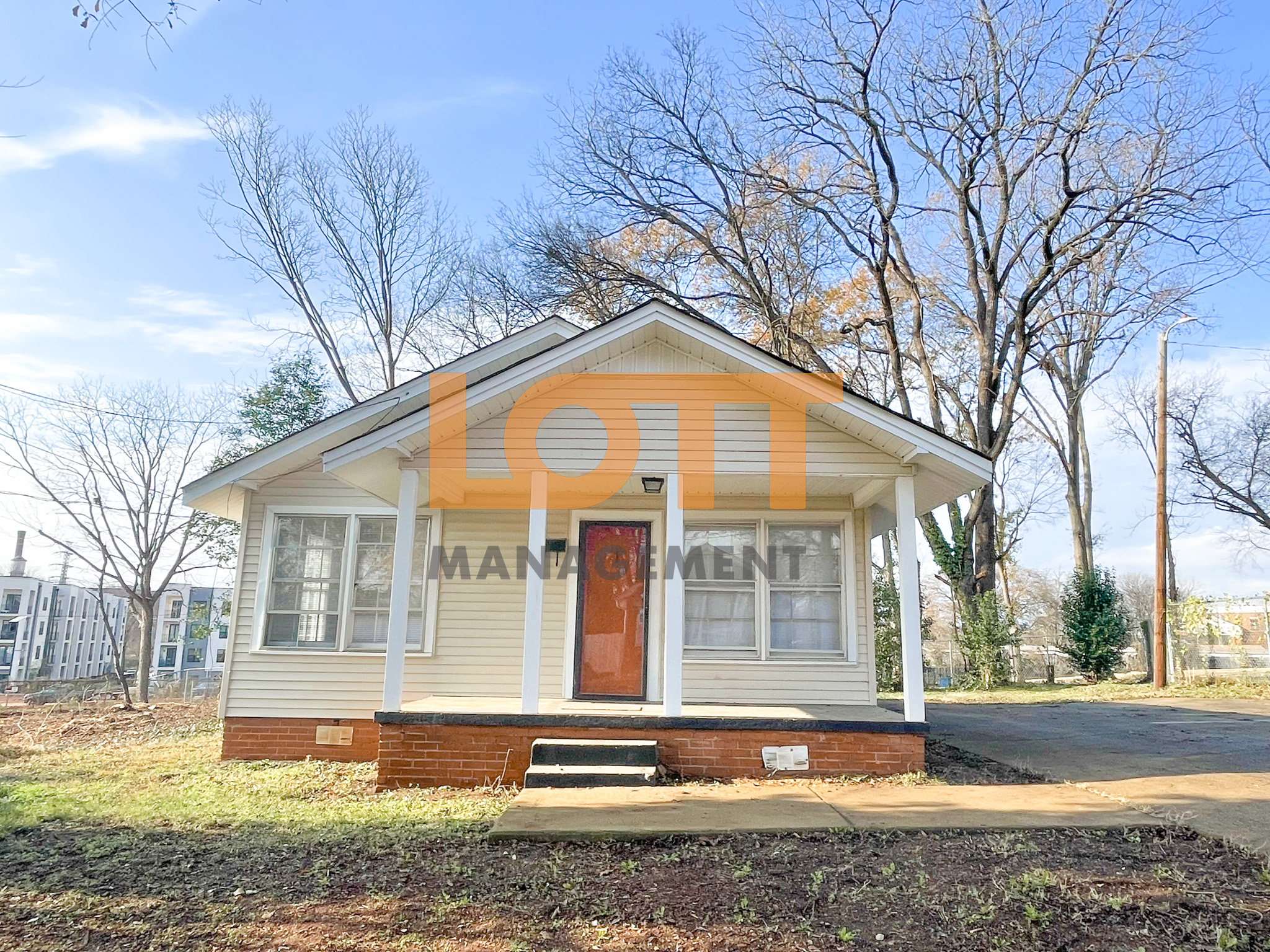 Front view of a single-story house with light-colored siding, a red brick foundation, a red front door, and a porch with white pillars. The yard features sparse grass and bare trees, with modern buildings visible in the background under a clear blue sky. A large semi-transparent 'LOTT MANAGEMENT' watermark overlays the image.