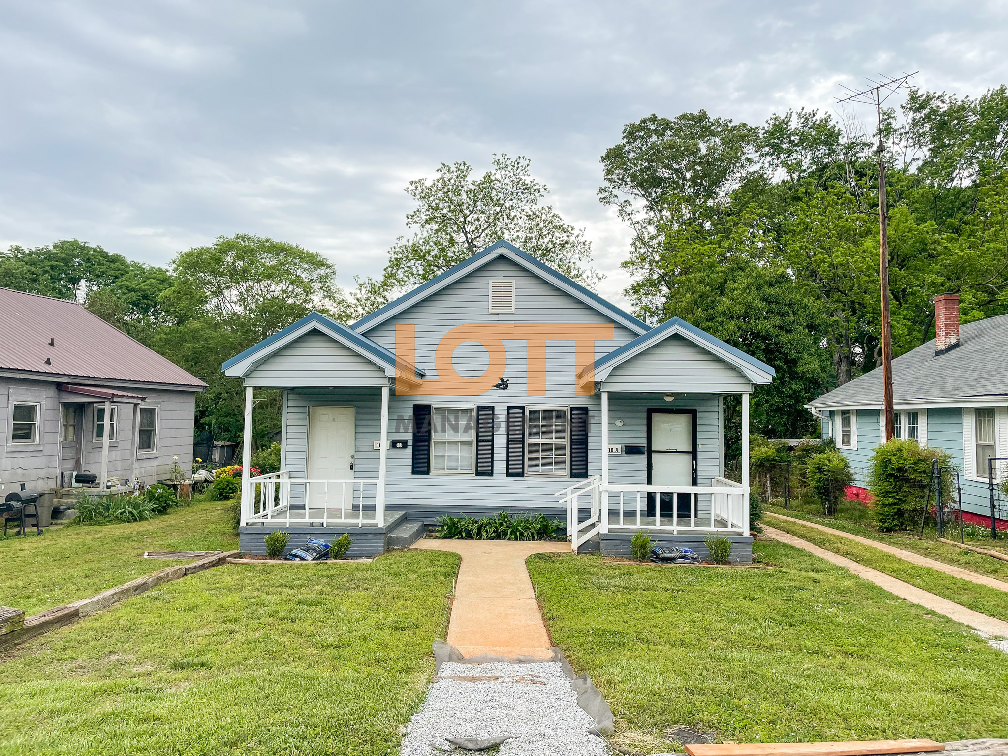Front view of a light blue, single-story duplex with two symmetrical porches, each supported by white square pillars and featuring white entry doors labeled '10 C' and '10 A.' The house has a gabled roof, black shutters, and a green lawn with a concrete walkway bordered by fresh gravel. Small bushes and landscaping materials are visible near the porches, indicating recent improvements. Neighboring houses and a utility pole are partially visible on either side, with a backdrop of tall green trees under an overcast sky. A watermark reading 'LOTT MANAGEMENT' overlays the image.