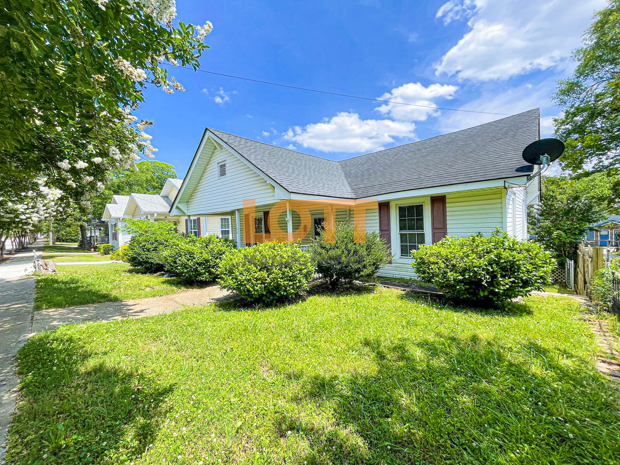 Wide view of a single-story house with light-colored siding, a dark gable roof, and reddish-brown shutters, surrounded by a green lawn and trimmed bushes. The house features a covered porch, a satellite dish, and a curved concrete pathway leading to the street. The background includes a tree-lined street with similar houses, under a bright blue sky with scattered white clouds. A semi-transparent watermark 'LOTT MANAGEMENT' overlays the house.