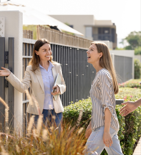 Two women outside a modern home entrance; one, a professional in a blazer holding a tablet, gestures invitingly, while the other, casually dressed, laughs as she walks forward. Lush landscaping and contemporary houses are visible in the sunny background.