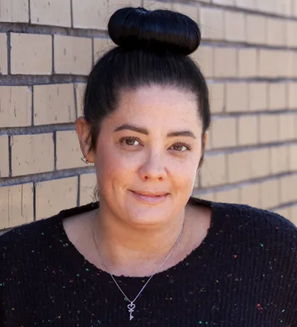 Portrait of Misty Rosen with dark hair in a high bun, wearing a black speckled sweater, a silver heart-shaped key pendant necklace, and small silver hoop earrings, set against a light brown brick wall.