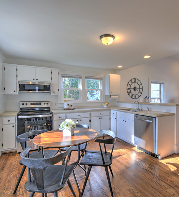 Bright kitchen and dining area with white Shaker cabinets, stainless steel appliances, light countertops, and warm hardwood floors. A round wooden dining table with black Windsor chairs and a white vase of flowers is centered in the foreground, with large windows providing natural light.