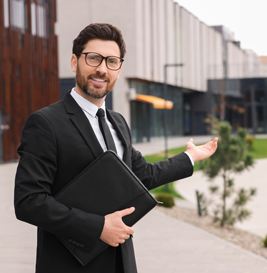 Smiling man in a black suit holding a folder, gesturing towards modern buildings with large glass windows, standing on a paved area with greenery in the background.