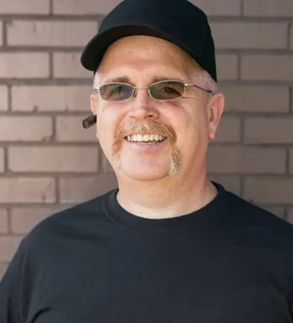 Portrait of Jim Crane. He's smiling. He has a handlebar mustache, wearing a black baseball cap, glasses, and a black t-shirt, with a small cigar tucked behind his ear, set against a light brick wall background.