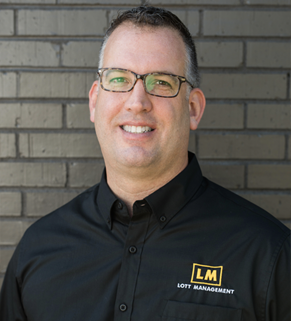 Professional portrait of Dan Lott. He is smiling. He has short dark hair and glasses, wearing a black shirt with a yellow 'LOTT MANAGEMENT' logo, set against a blurred dark brick wall background.
