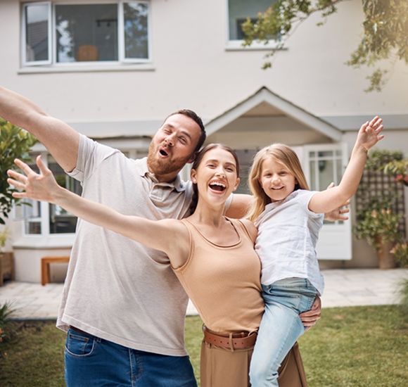 Joyful family of three—a father, mother, and young daughter—smiling with outstretched arms in their backyard, standing in front of a light-colored two-story house with a green lawn and lush trees on a sunny day.