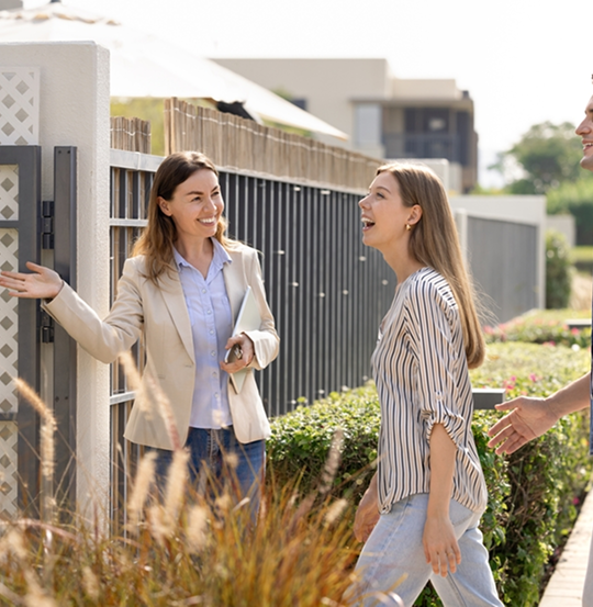 Smiling female agent holding a tablet and keys, gesturing towards an open gate while engaging with a laughing woman and her companion, set in a modern residential area with manicured greenery and contemporary buildings in the background.
