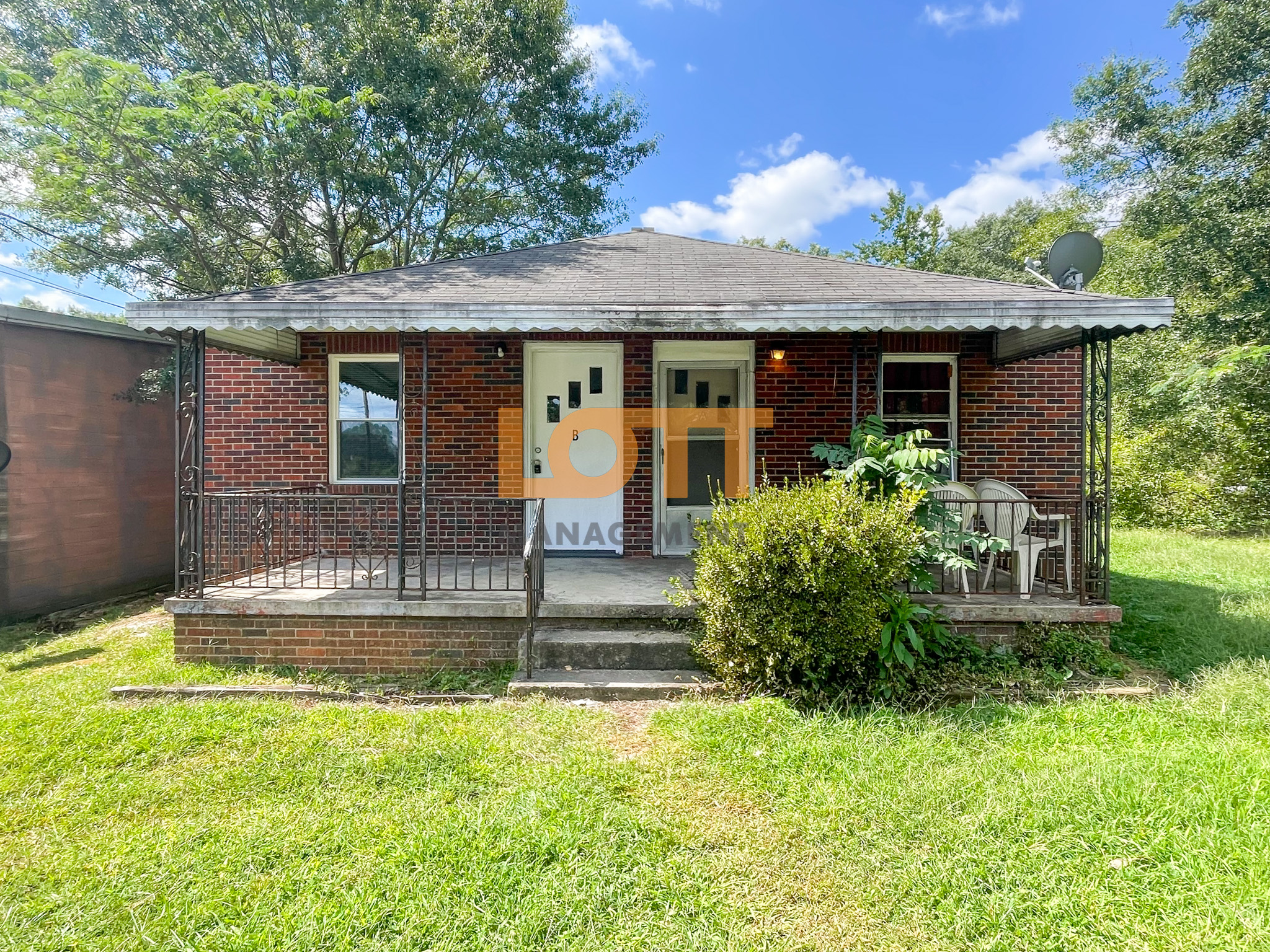 Single-story red brick house with a dark gray shingled roof, a front porch covered by a white corrugated metal awning, and decorative metal railings. The porch features two doors, outdoor chairs, and green plants. Surrounded by a green lawn, tall trees, and a bright blue sky. A semi-transparent 
